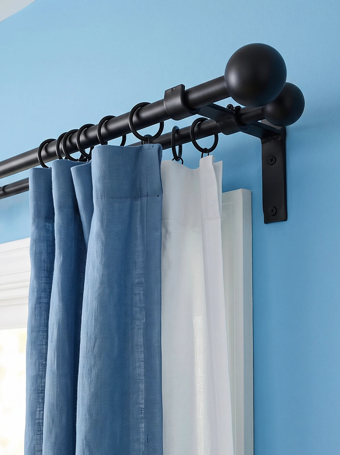 Black metal curtain rod with decorative finials and wall bracket mounted on a light blue wall, holding blue and white curtains.