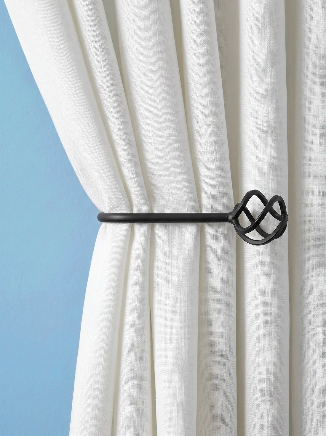 A close-up of a white curtain tied back with a black metal curtain rod featuring an ornate knot-like finial, set against a light blue background.
